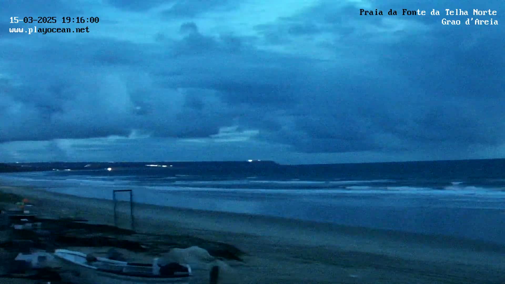 A dark blue sky with clouds hangs over a beach at twilight, where small waves lap the shore near some boats.