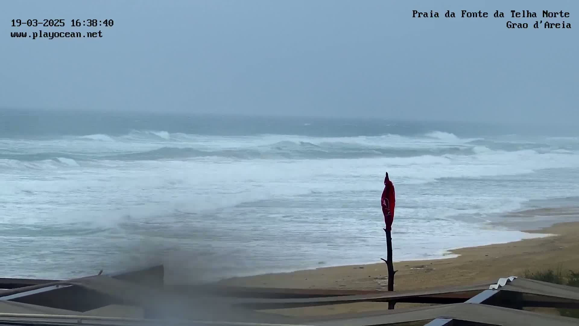 A stormy ocean with choppy waves crashes onto a sandy beach, where a red flag stands on a post.