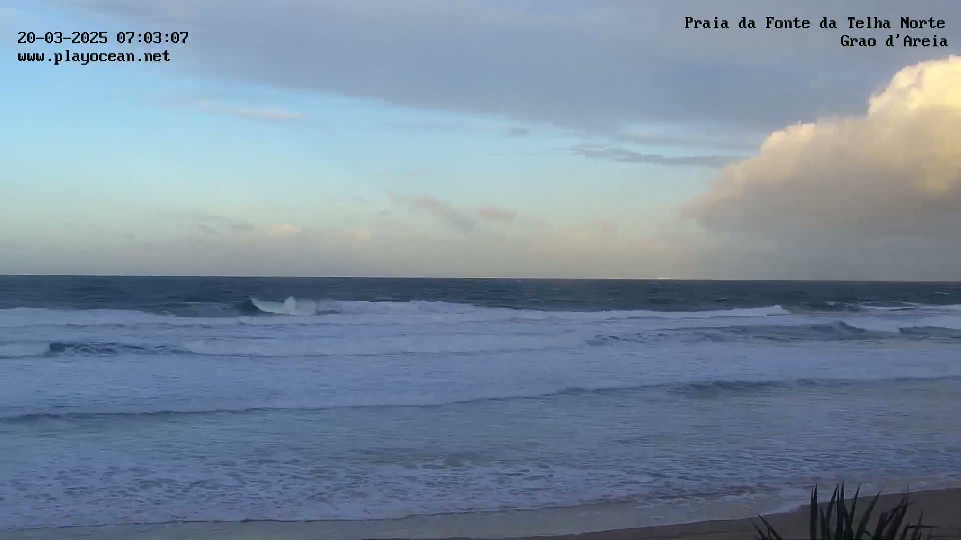 A mostly cloudy sky overlooks a beach with moderate waves rolling onto the shore.