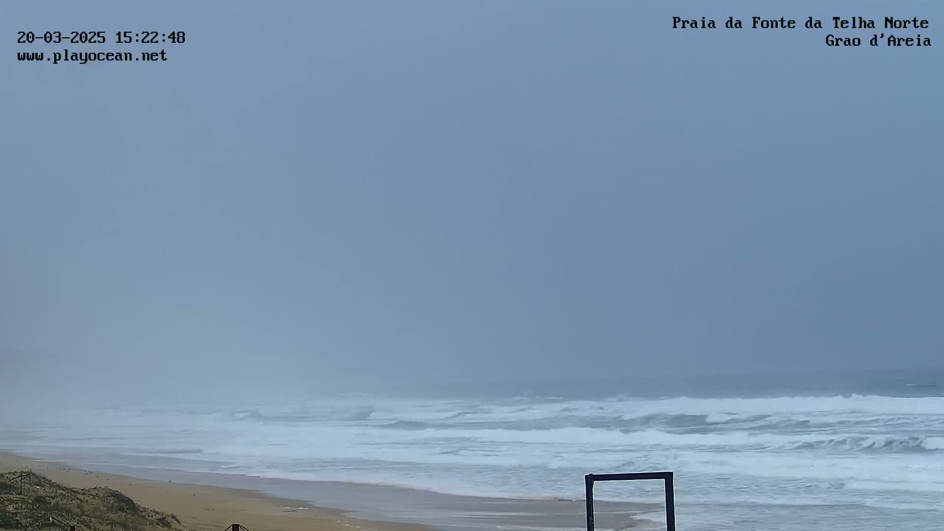 A hazy, overcast day shows a sandy beach with moderate waves crashing on the shore.