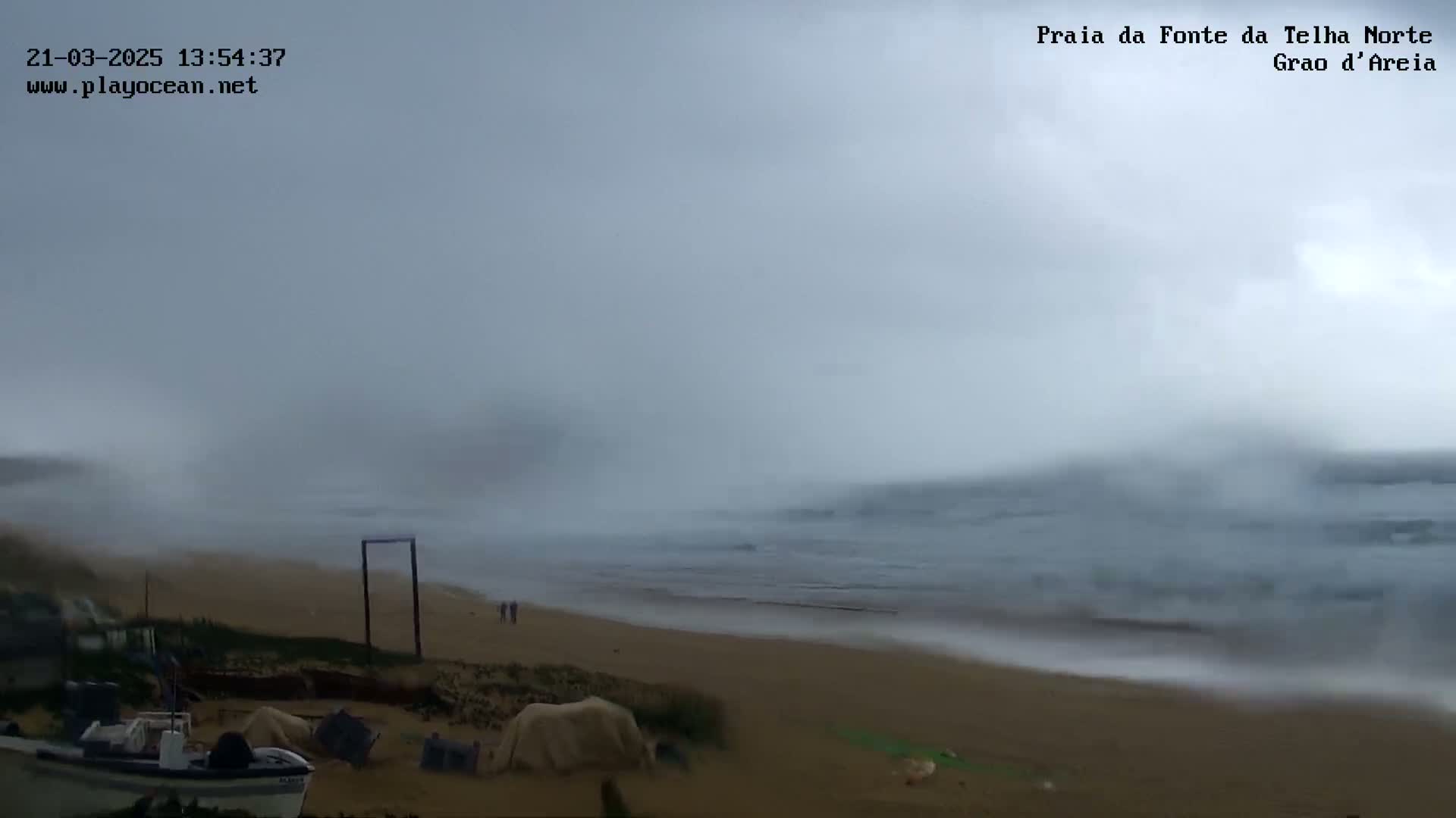 A mostly overcast day shows a beach with a few people, some debris and a small boat near the shore, and somewhat choppy ocean waves.