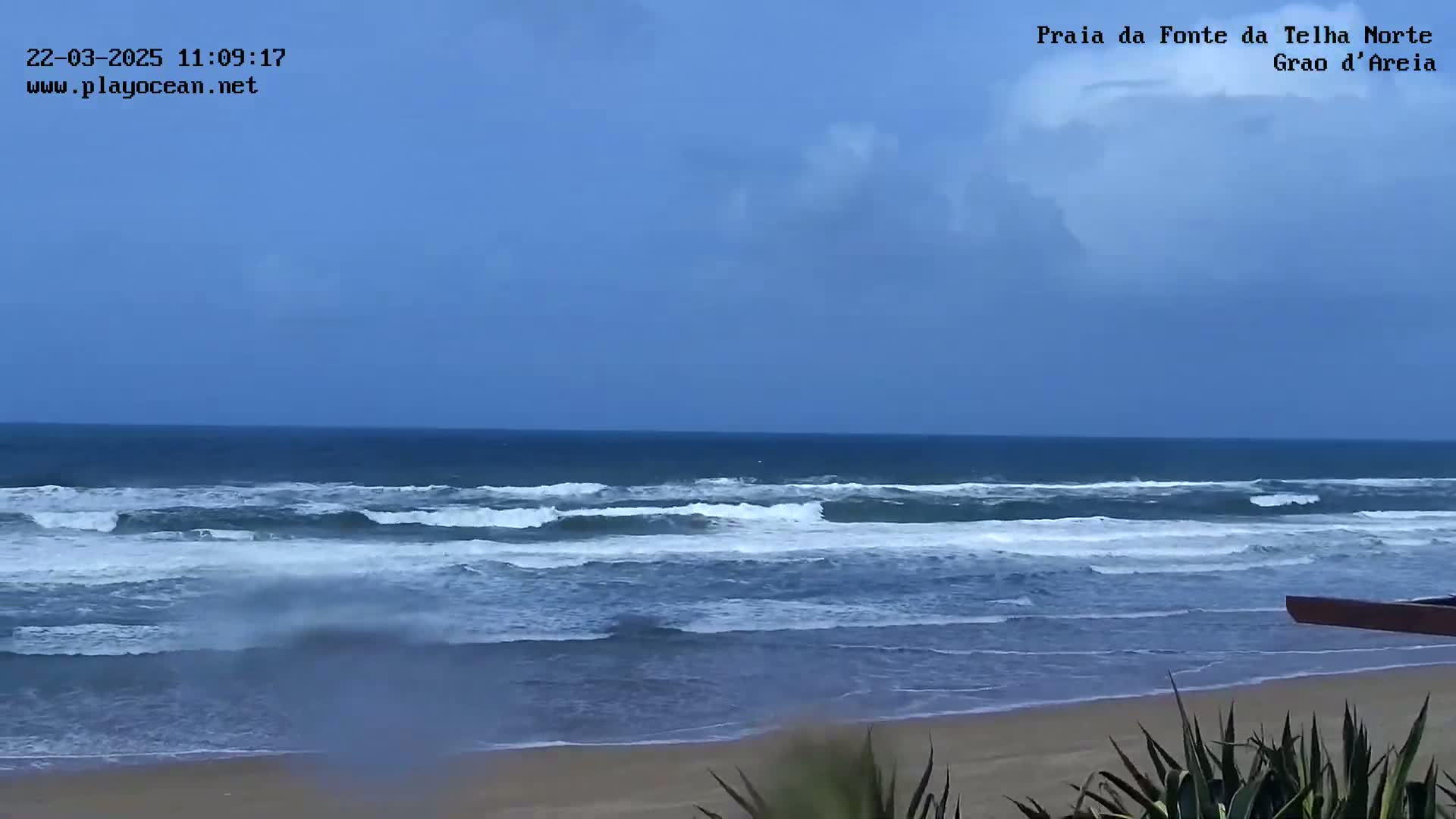 A partly cloudy day shows a wide sandy beach with moderate waves rolling into the shore.