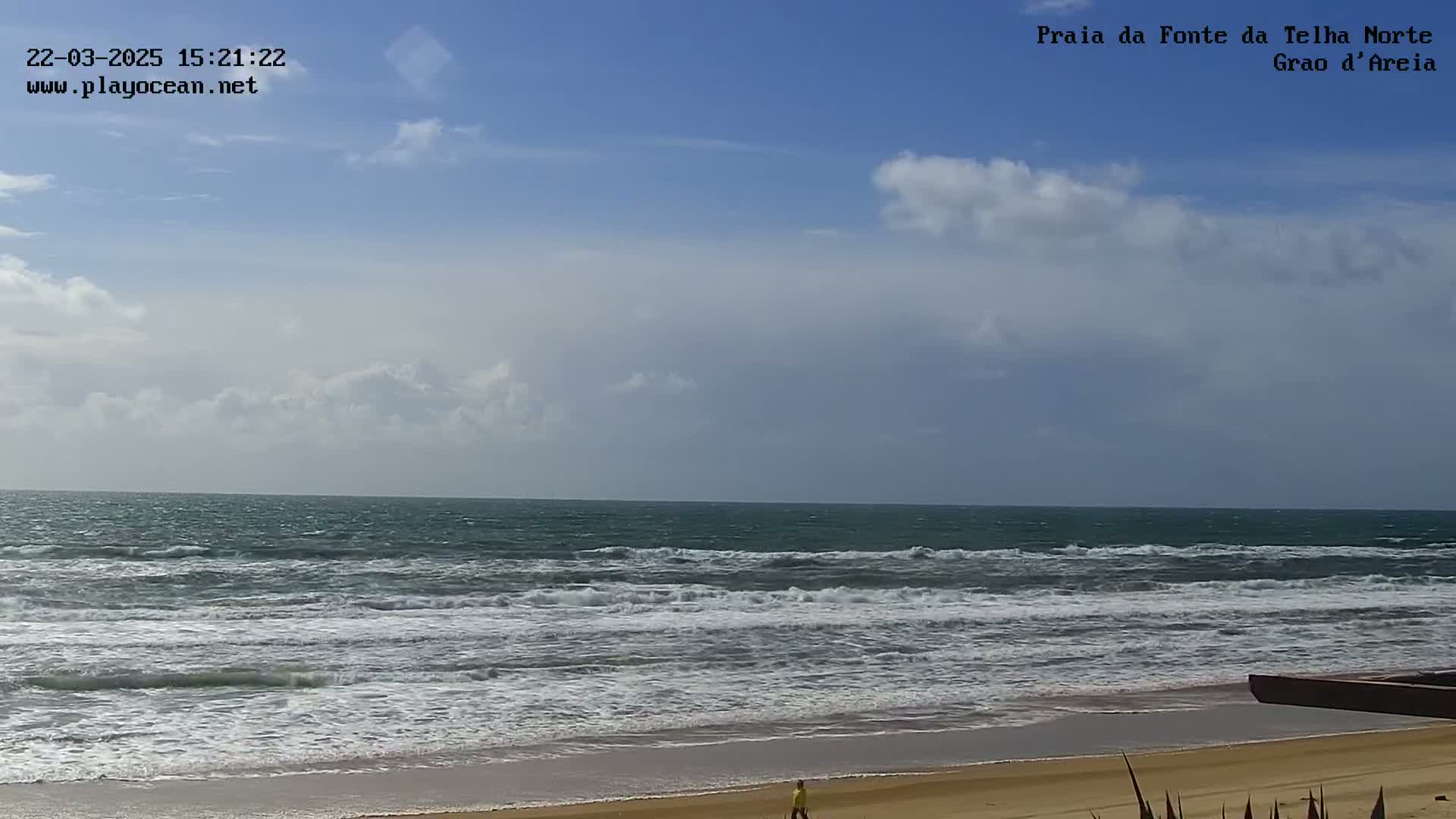 A mostly cloudy sky overlooks a sandy beach with moderate waves breaking on the shore, and a lone person walking on the sand.