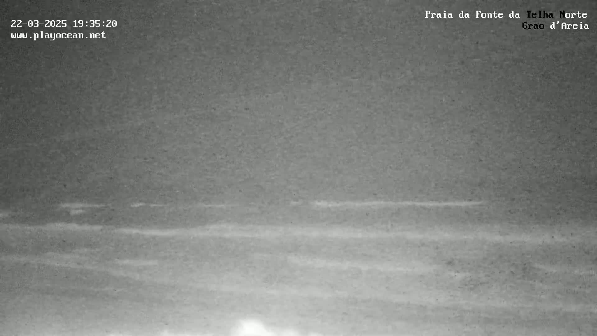 A nighttime, low-light image shows a dark, mostly calm ocean with faint waves visible near the shore.