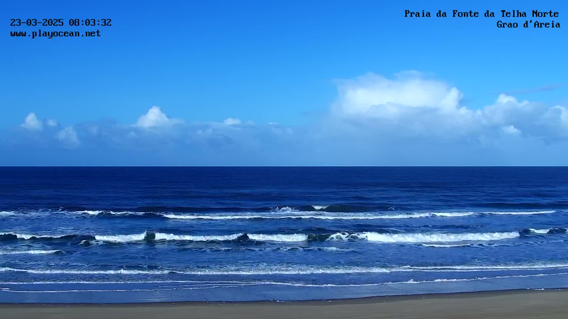 Under a mostly clear blue sky with a few cumulus clouds, gentle waves roll onto a sandy beach.