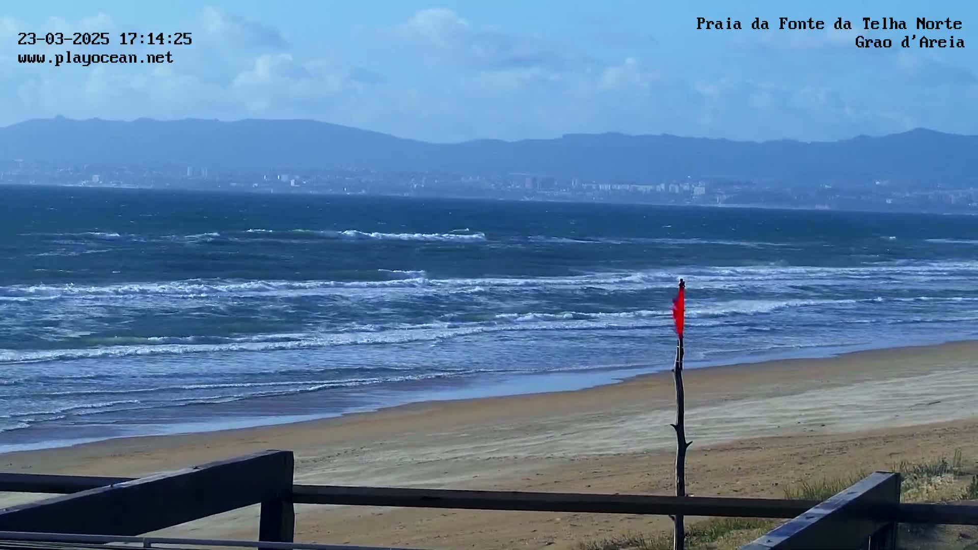 A sandy beach with moderate waves and a distant city under a partly cloudy sky.