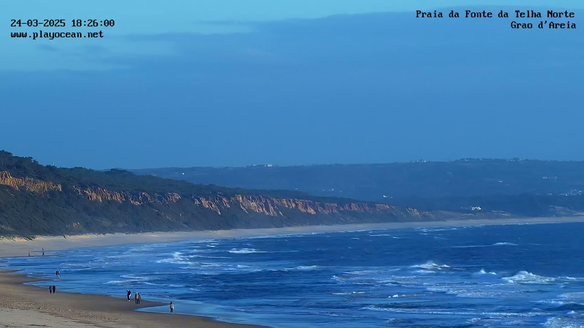 A sandy beach with a few people walking along the shore stretches beneath a cliff covered in dark green vegetation, meeting a blue ocean with moderate waves under a clear sky.