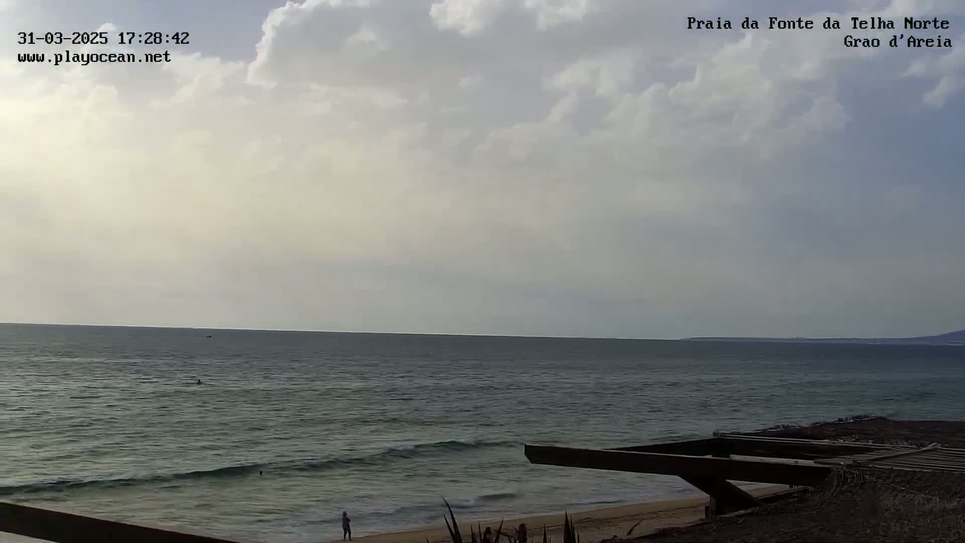 A mostly cloudy sky overlooks a calm ocean with small waves lapping a sandy shore, where a person stands near a partially constructed wooden structure.