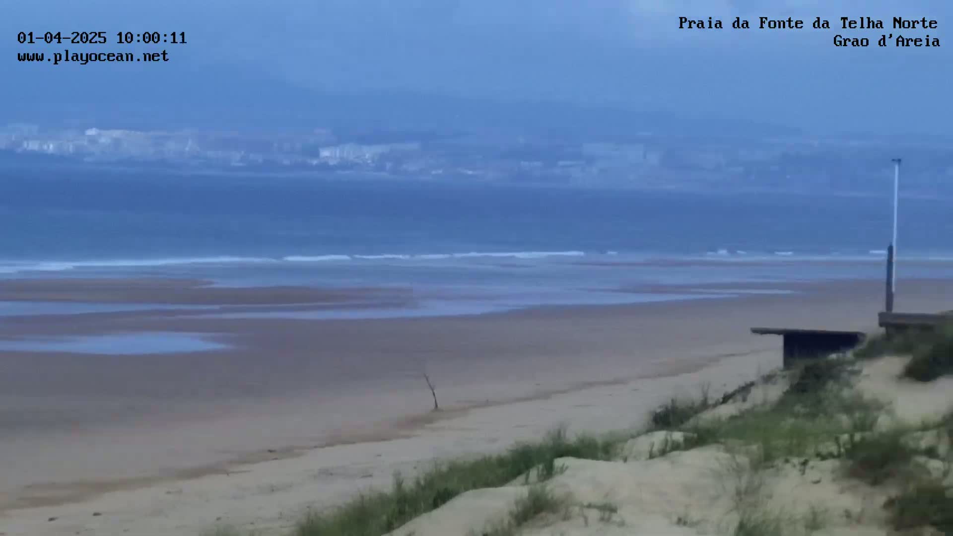 A wide sandy beach with small waves and a hazy, overcast sky stretches towards a distant town.