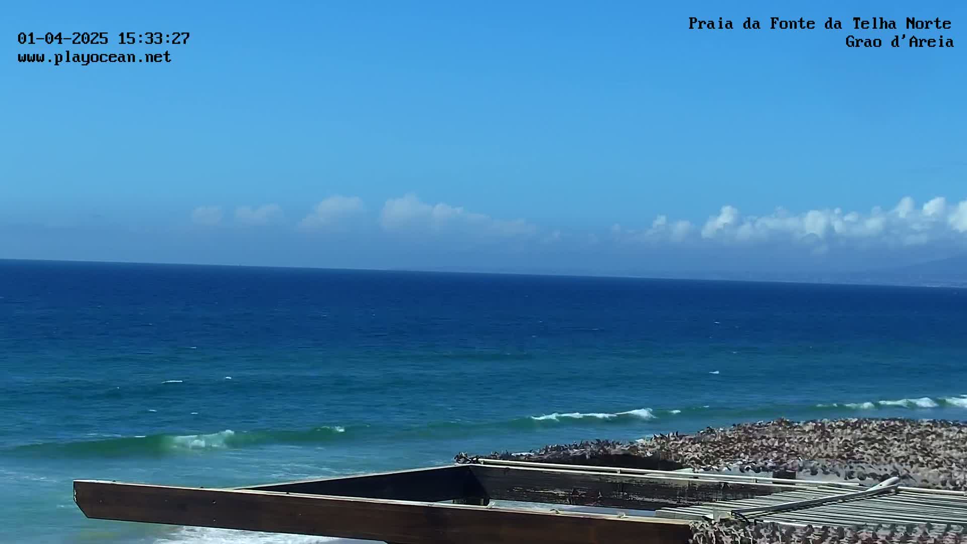 A mostly clear blue sky overlooks a blue ocean with small waves breaking on a rocky shore, partially obscured by a wooden structure in the foreground.
