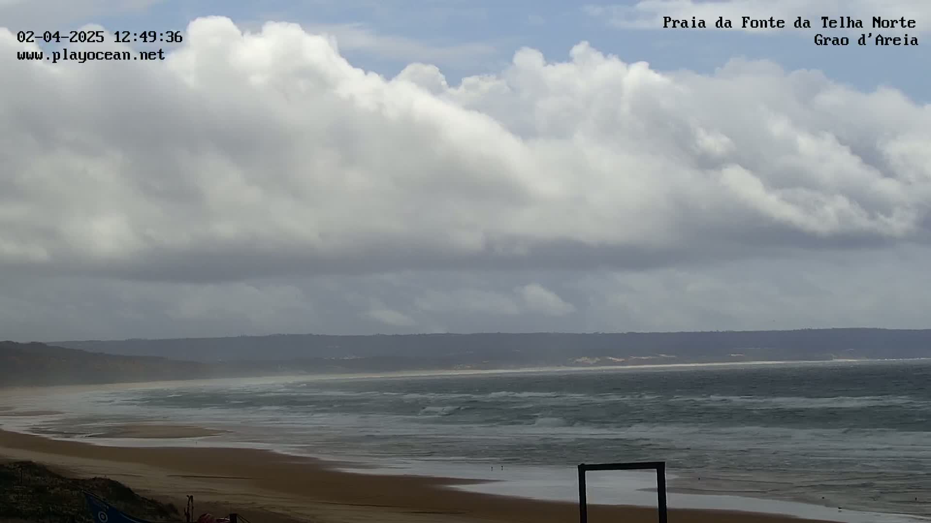 A mostly cloudy sky hangs over a wide, sandy beach with moderate waves lapping the shore.