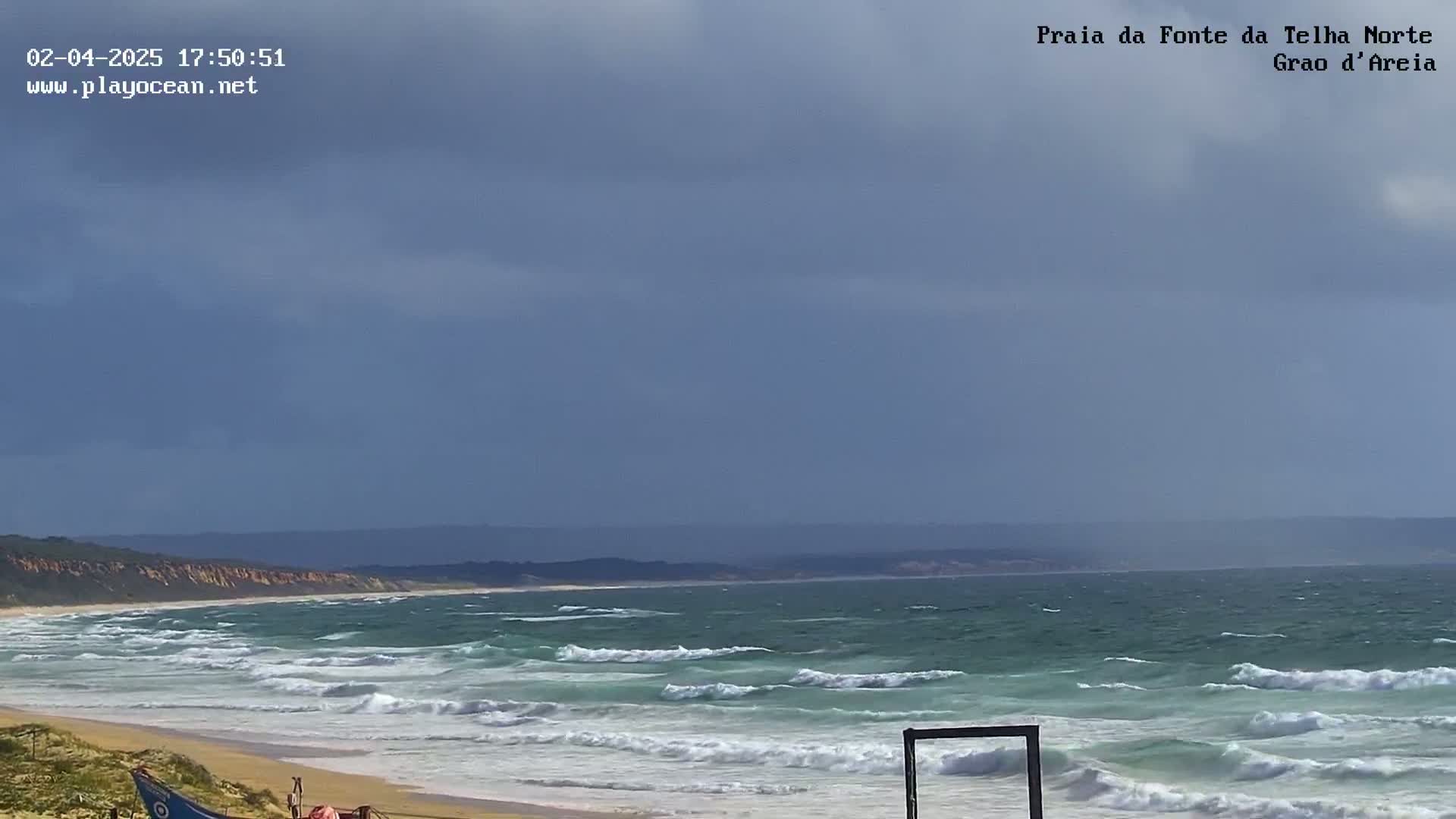 A sandy beach with moderate waves under a mostly cloudy sky meets a teal ocean, backed by a distant shoreline.