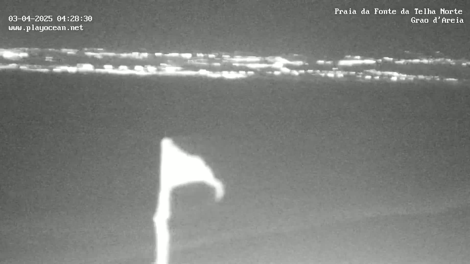 A nighttime, low-light view of a dark body of water with a faint line of city lights in the distance and a flagpole with a flag blowing in the wind in the foreground.