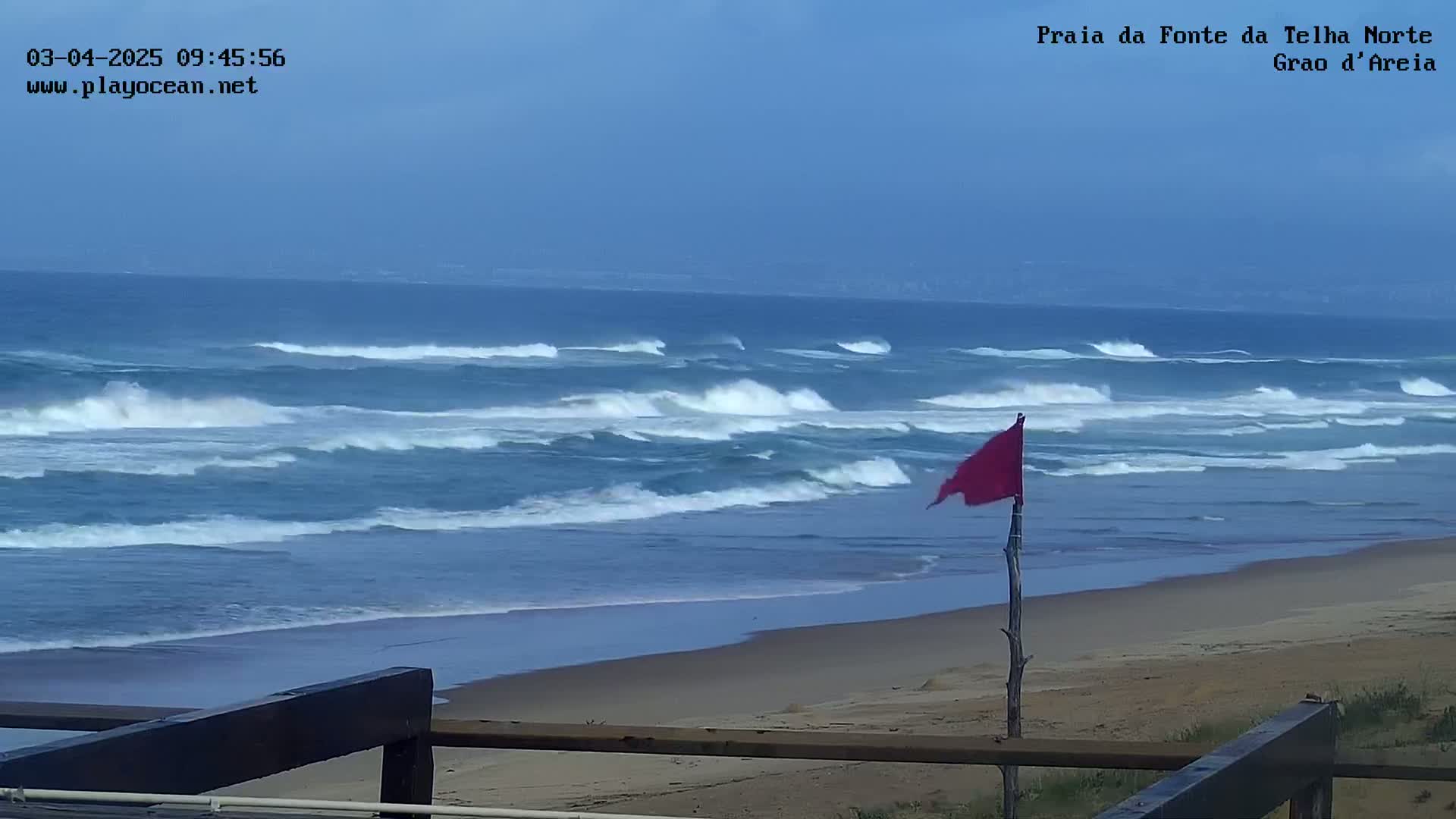 A mostly cloudy day shows moderate waves crashing on a sandy beach with a red flag on a post indicating caution.