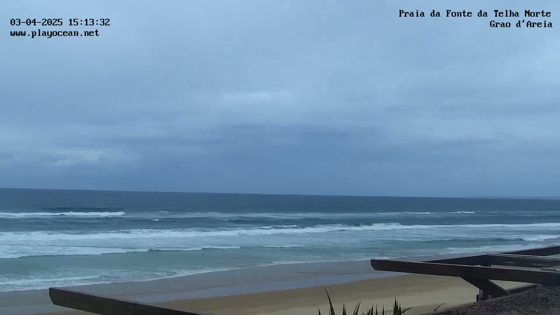A mostly cloudy sky overlooks a beach with gentle waves rolling onto the shore, partially obscured by a wooden structure in the foreground.