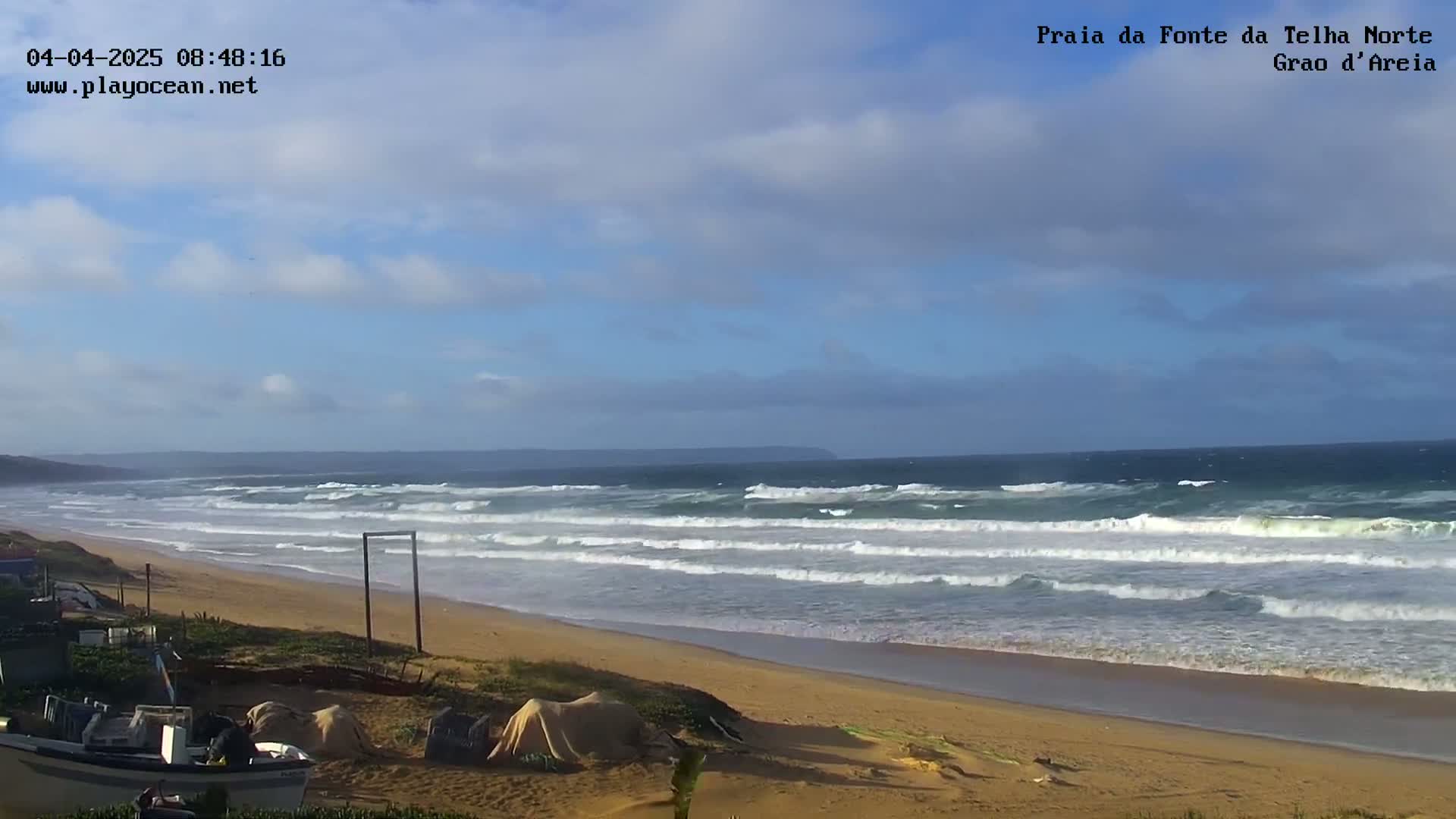 A sandy beach with several small boats on the shore is met by a partly cloudy sky and a dark blue ocean with moderate waves.