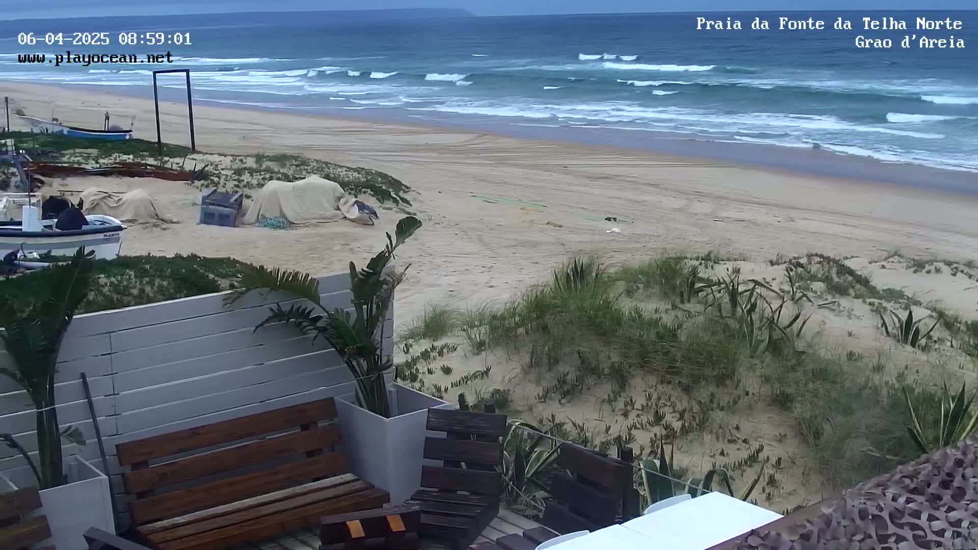 A sandy beach with moderate waves under an overcast sky is viewed from a patio with wooden furniture and potted plants.