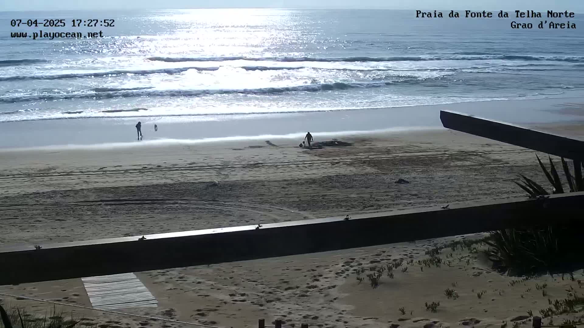 A sandy beach with gentle waves under a bright, sunny sky shows a few people walking near the water's edge.