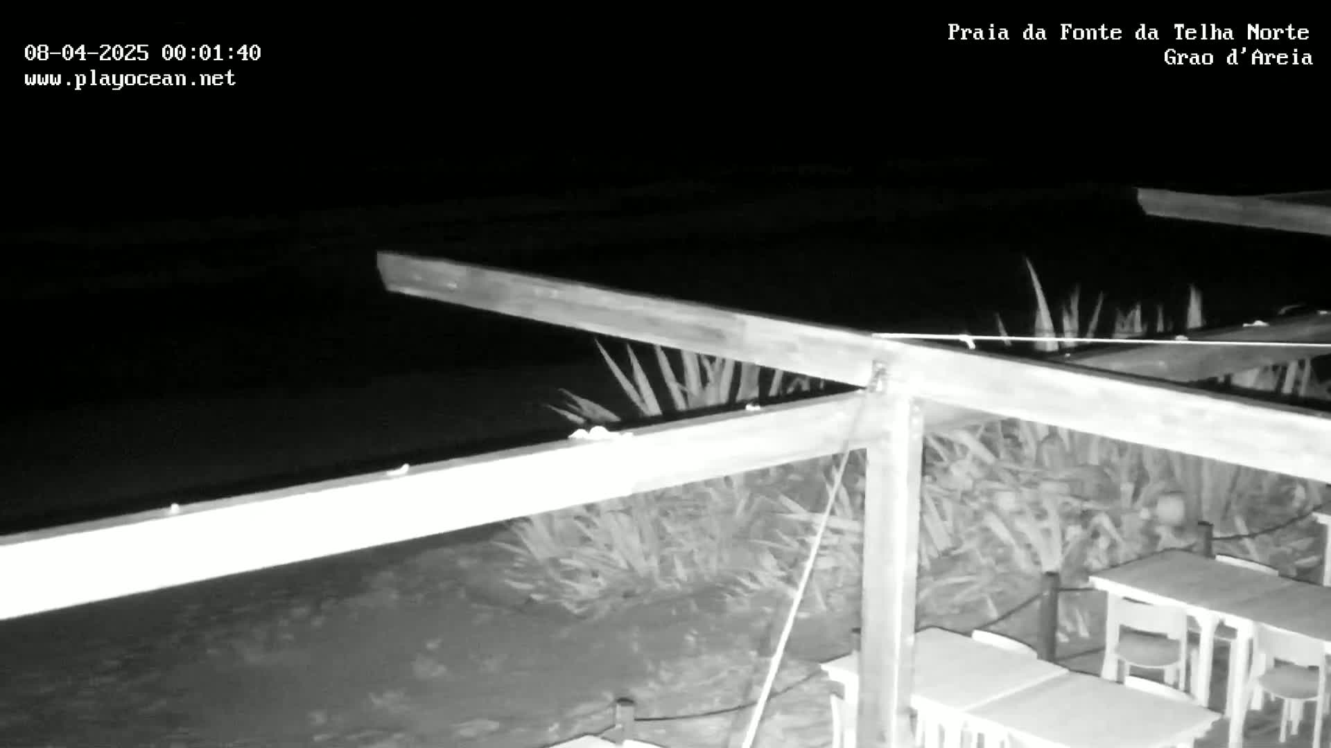 A nighttime, black-and-white image shows a wooden pergola structure overlooking a dark body of water, with several empty tables and chairs situated below.