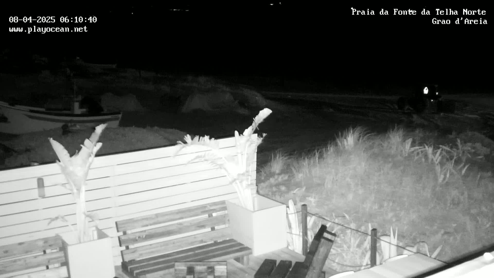 A nighttime black-and-white image shows a beach scene with a wooden fence and potted plants in the foreground, a vehicle in the midground, and a boat and dark ocean in the background.