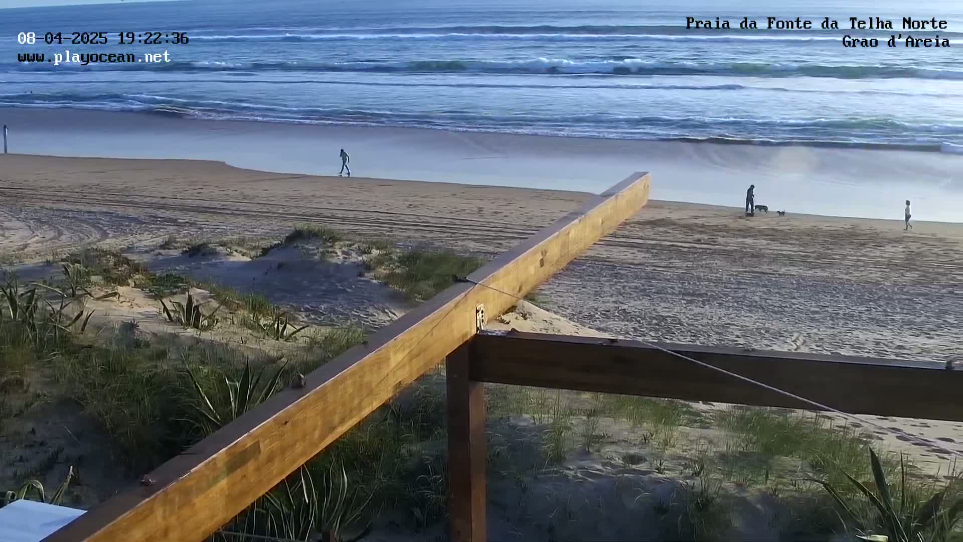 A calm ocean with small waves laps a sandy beach where a few people stroll, viewed from behind a wooden structure overlooking the scene.