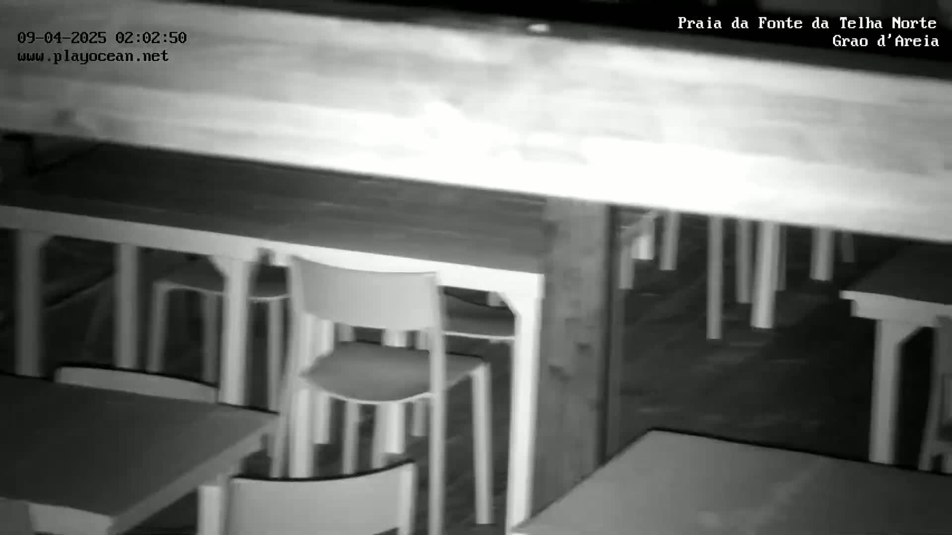 A low-light grayscale image shows numerous empty chairs and tables arranged under a structure at night.