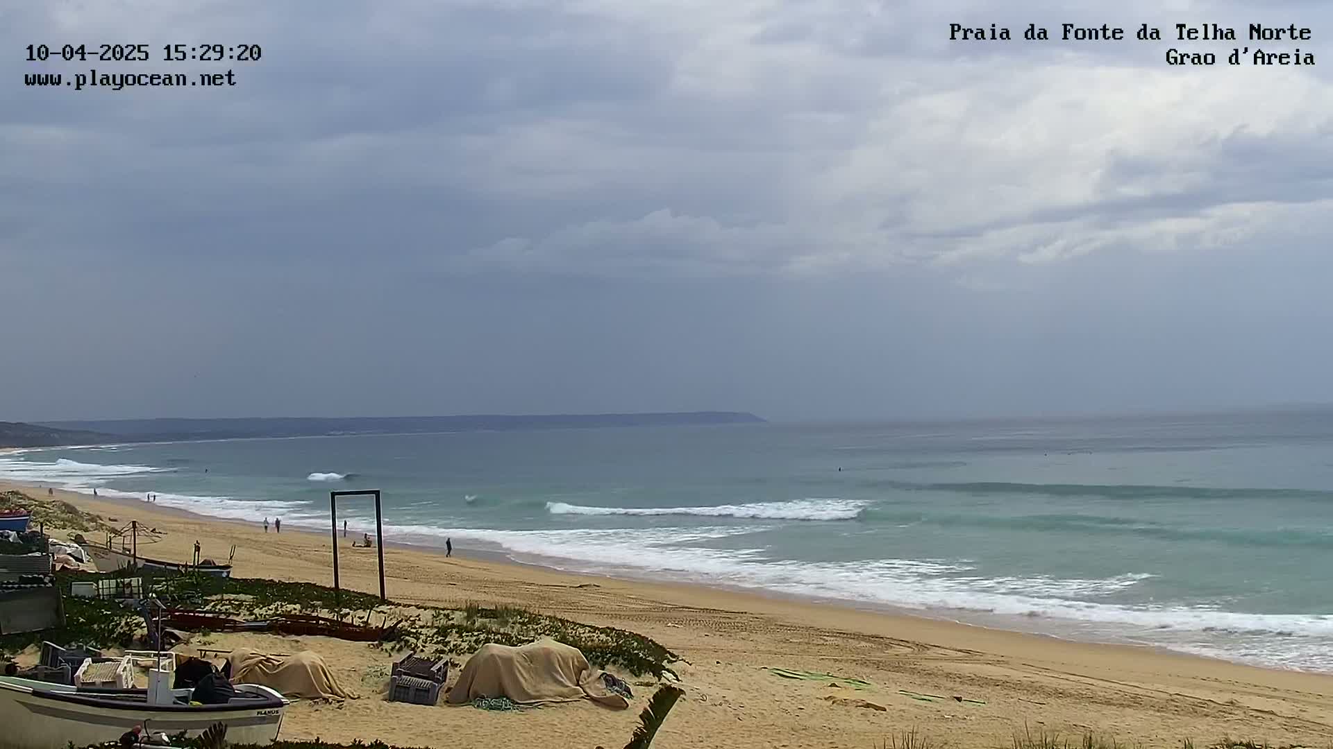 A sandy beach with gentle waves and a few people on it stretches out under a cloudy sky, next to some fishing boats and equipment.