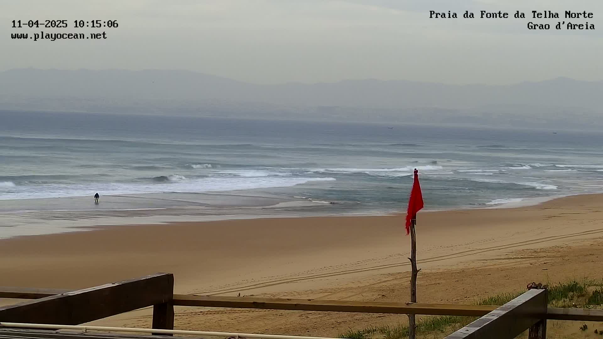 A sandy beach with small waves under an overcast sky, a single person walking on the wet sand, and a red flag on a stick in the sand.