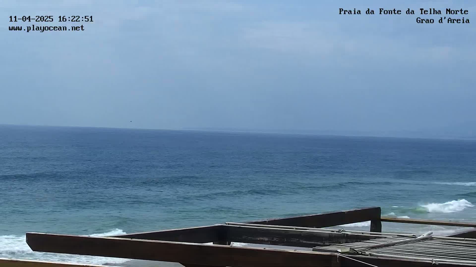A mostly clear sky overlooks a calm ocean with small waves breaking on a sandy shore, partially obscured by a dark wooden structure.