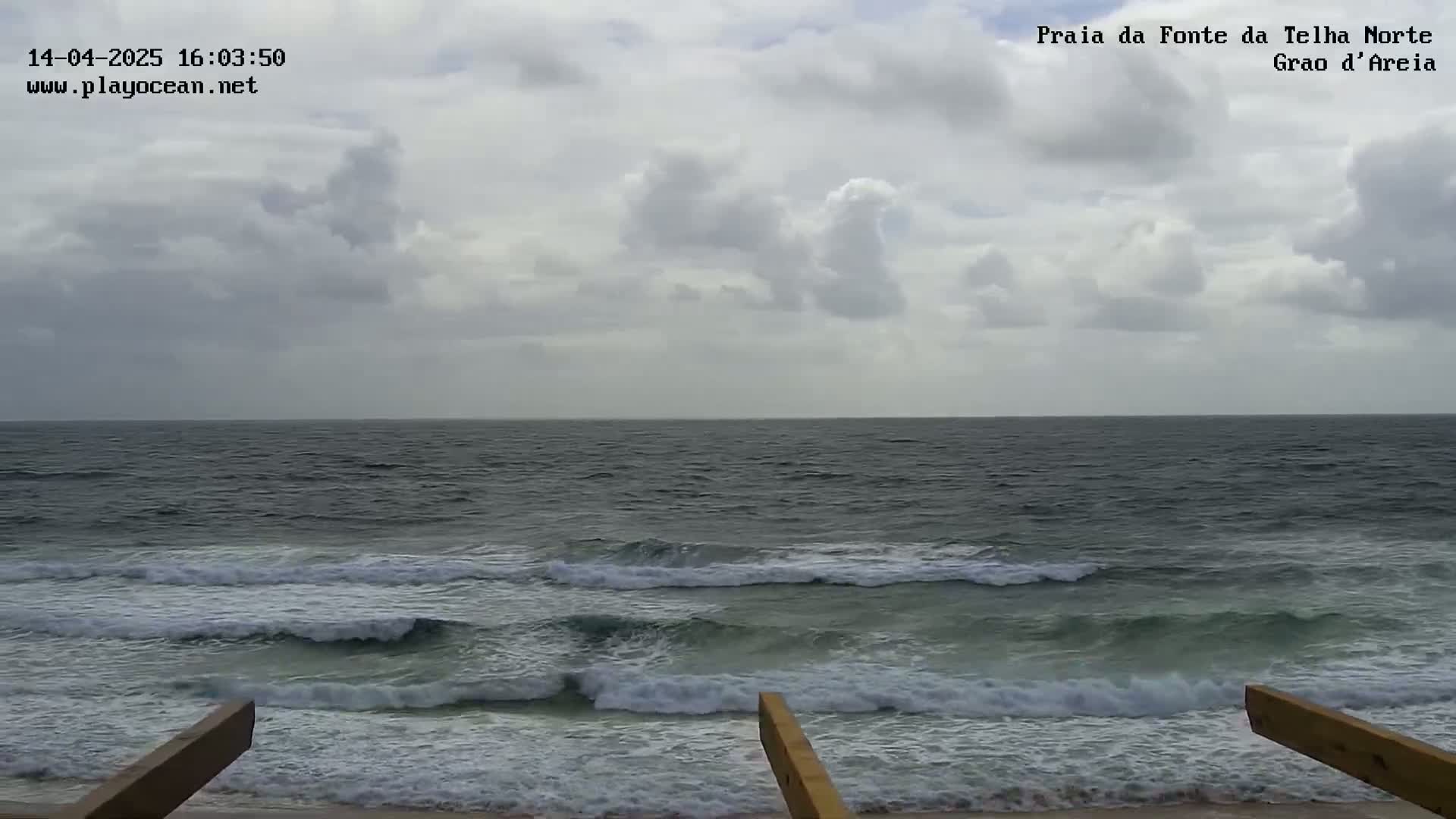 An overcast sky hangs over a dark-grey ocean with moderate waves breaking on a sandy shore, partially obscured by wooden beams in the foreground.
