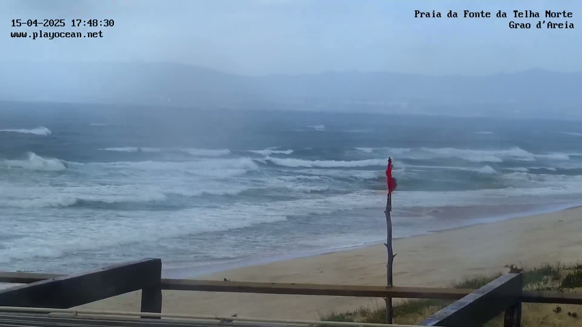 A beach with choppy waves under an overcast sky is viewed from behind a wooden railing.
