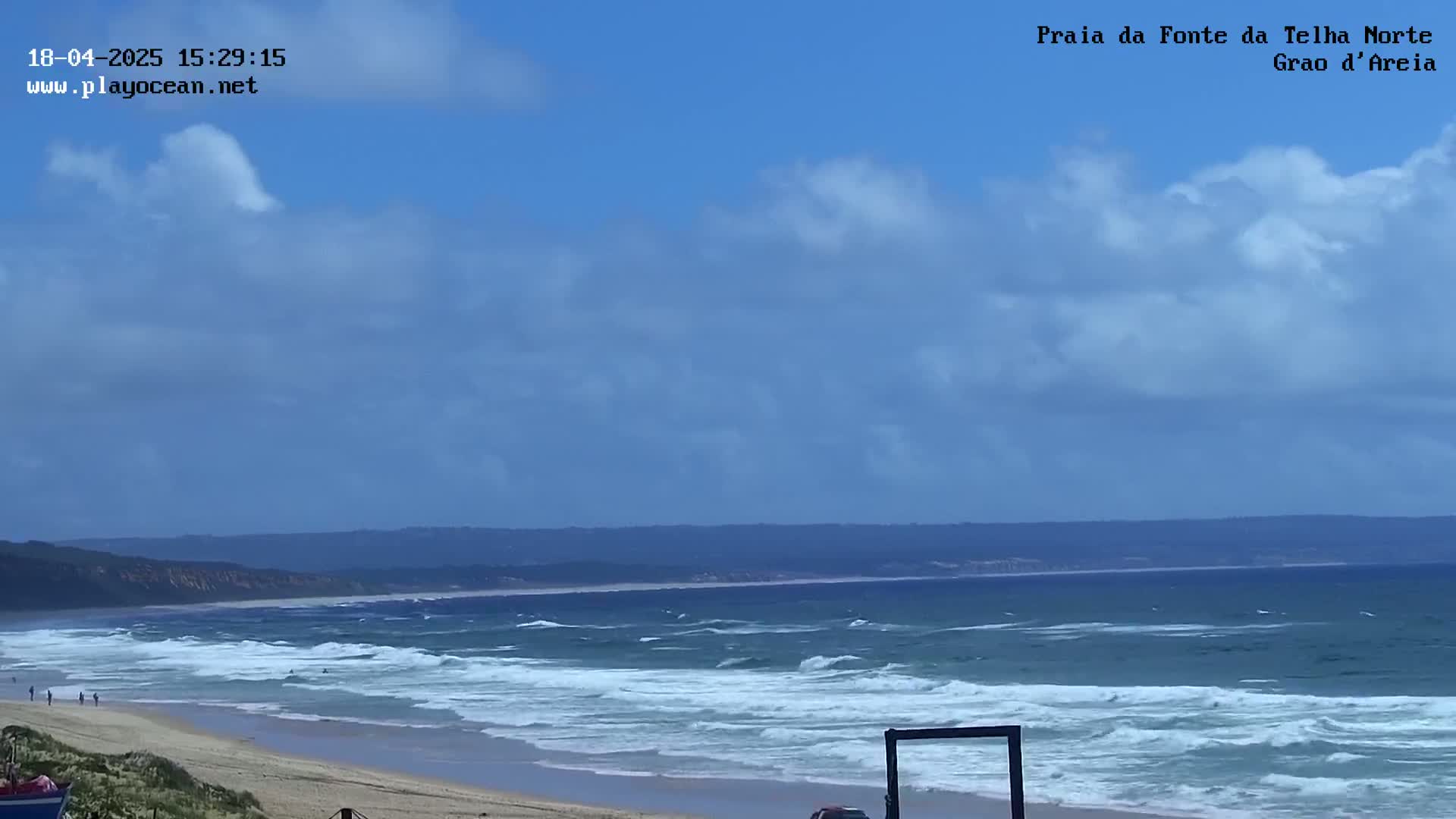 A sandy beach with moderate waves under a partly cloudy sky stretches to a distant coastline.
