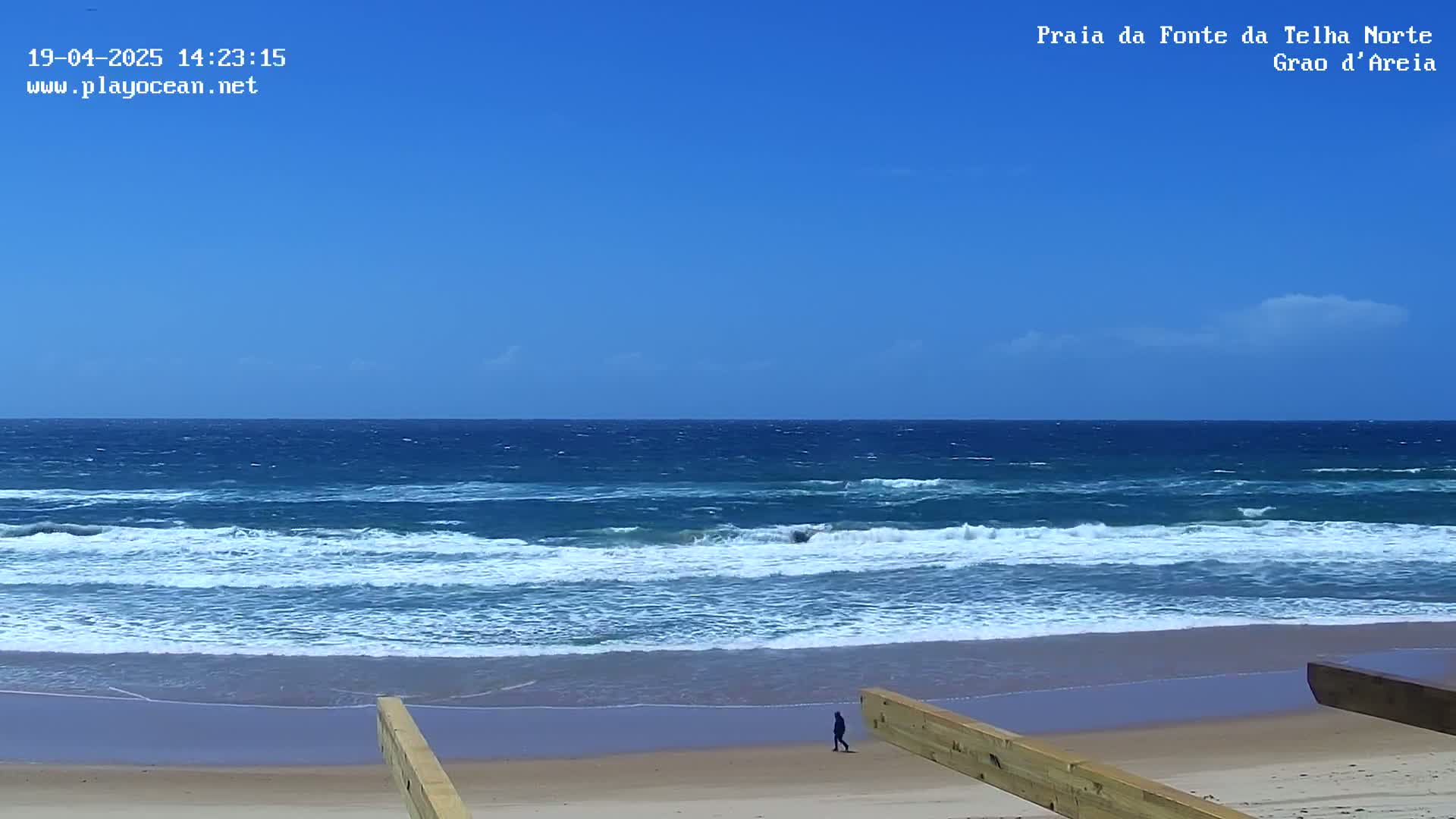 A person walks on a sandy beach under a clear blue sky, with moderate waves breaking on the shore.
