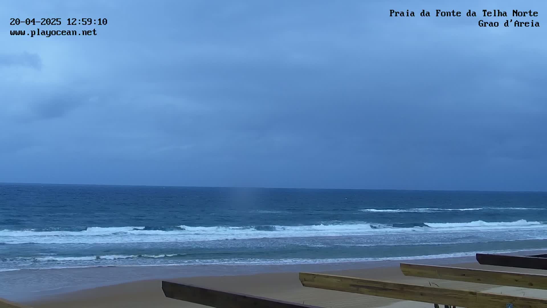 Under a mostly cloudy sky, waves roll onto a sandy beach with wooden structures in the foreground.