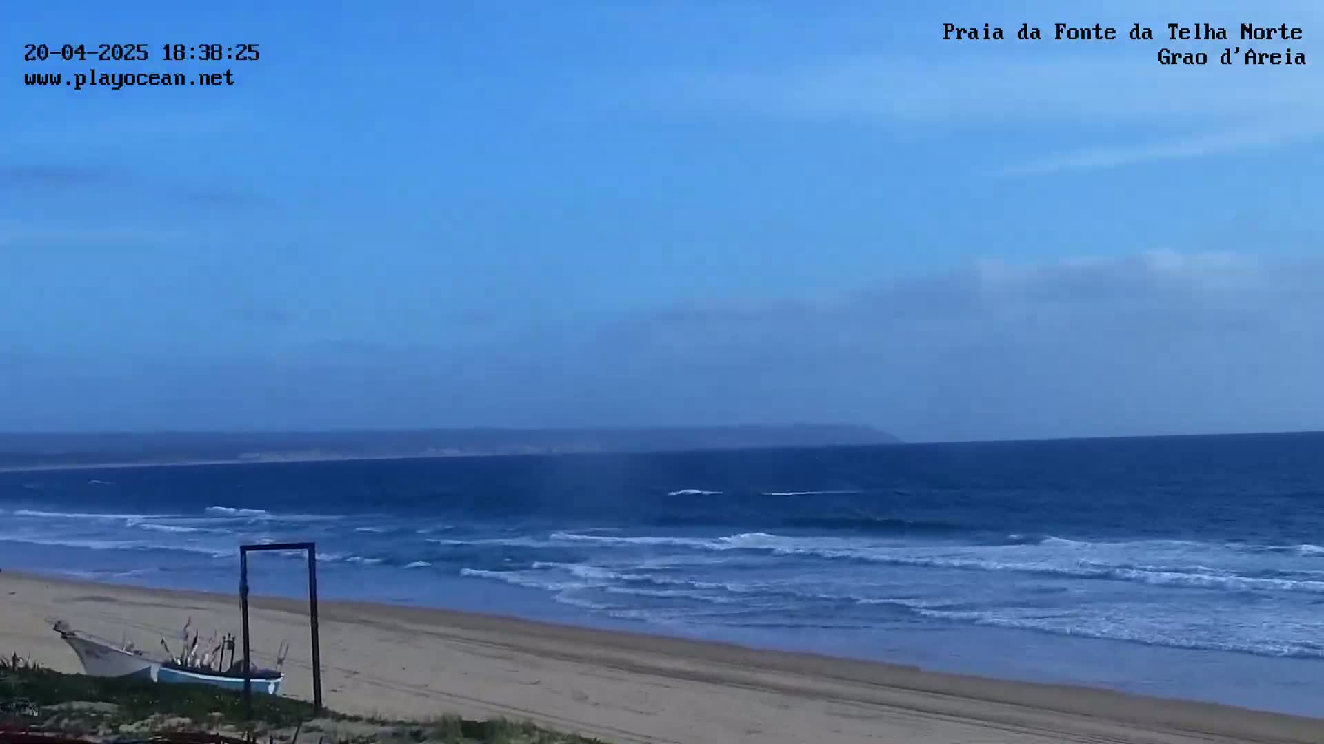 A sandy beach with gentle waves under a clear blue sky meets a calm ocean with a distant shoreline.