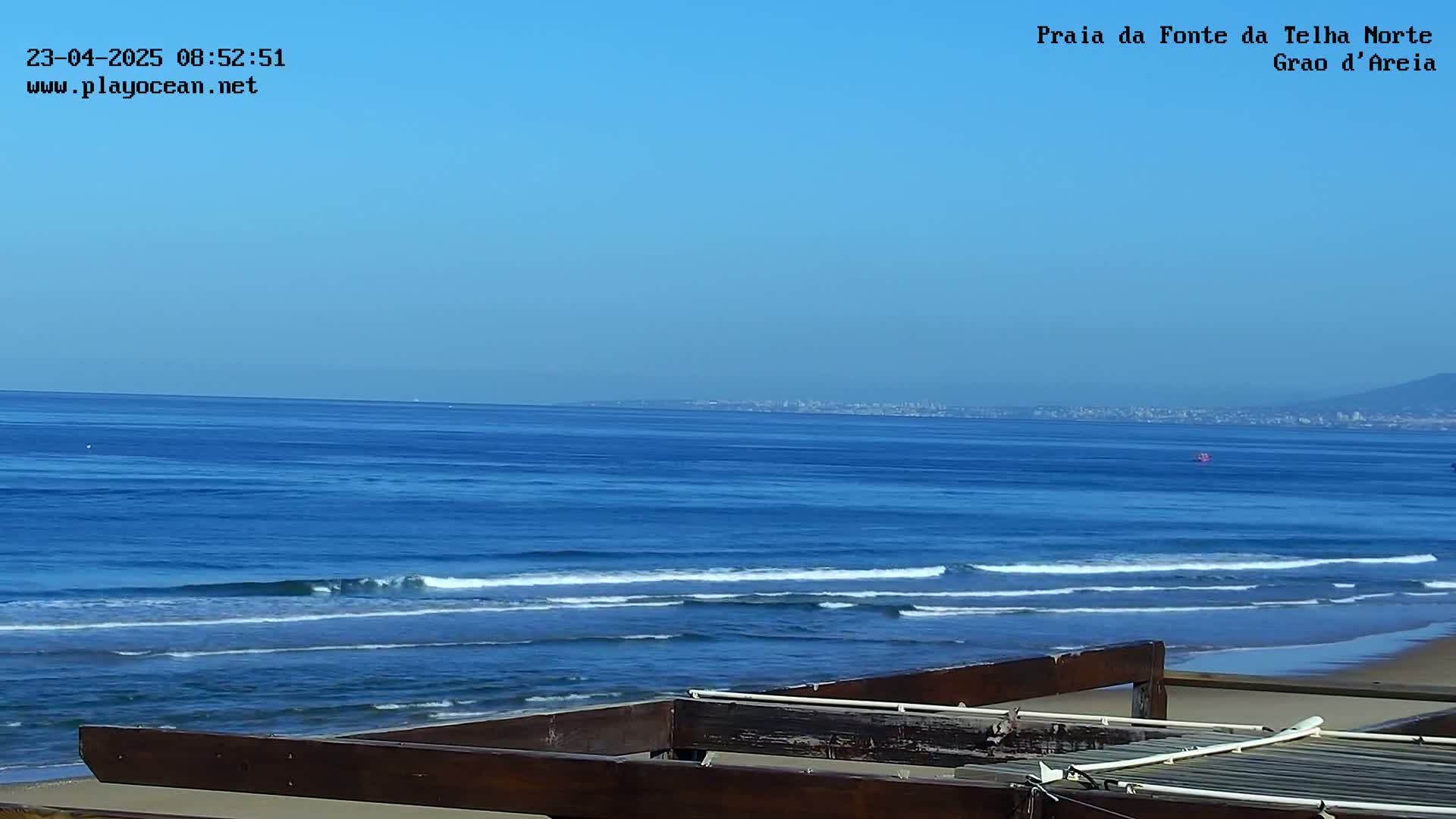 A tranquil ocean scene under a clear blue sky shows a sandy beach with gentle waves rolling onto the shore, partially obscured by a wooden structure in the foreground.