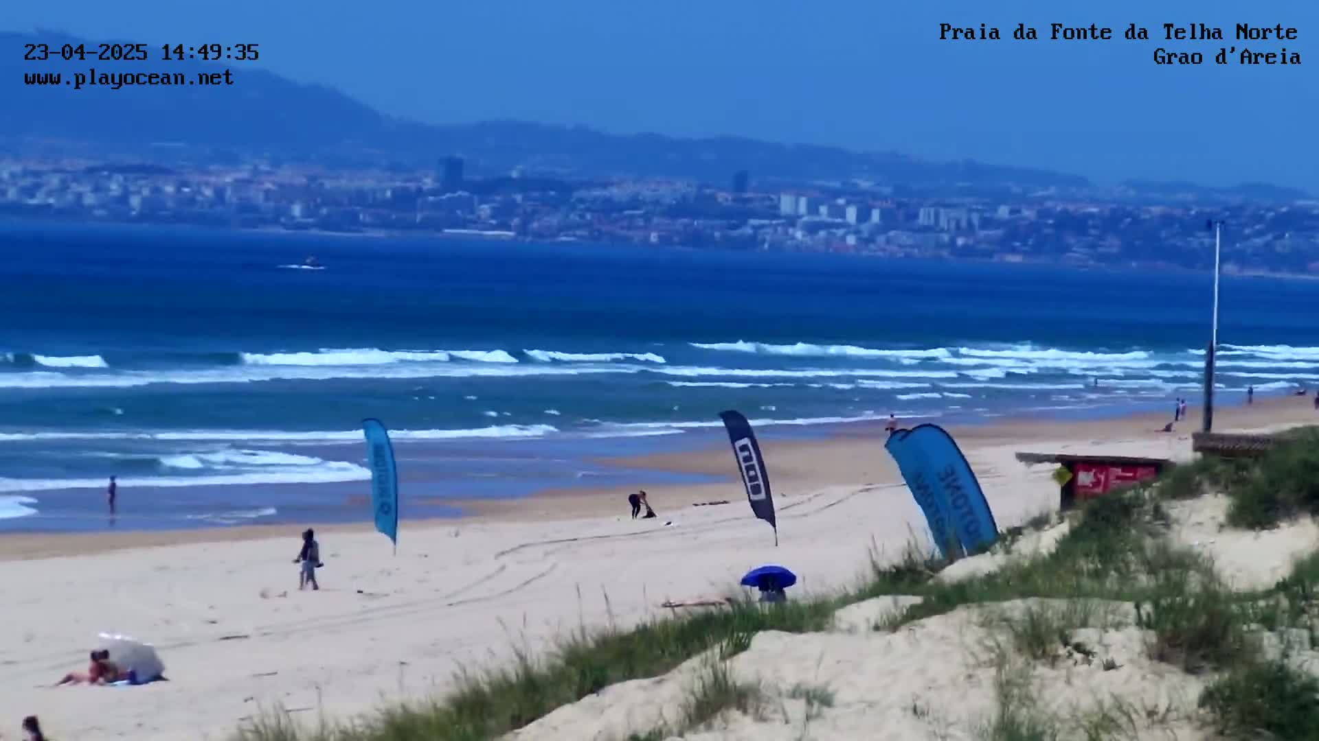 A sandy beach with a few people and several flags is visible, overlooking a calm ocean with gentle waves and a distant city skyline under a clear sky.