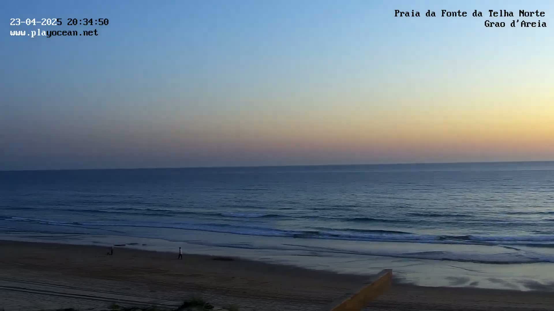 A tranquil beach scene at sunset shows gentle waves lapping a sandy shore with a few people walking in the distance.