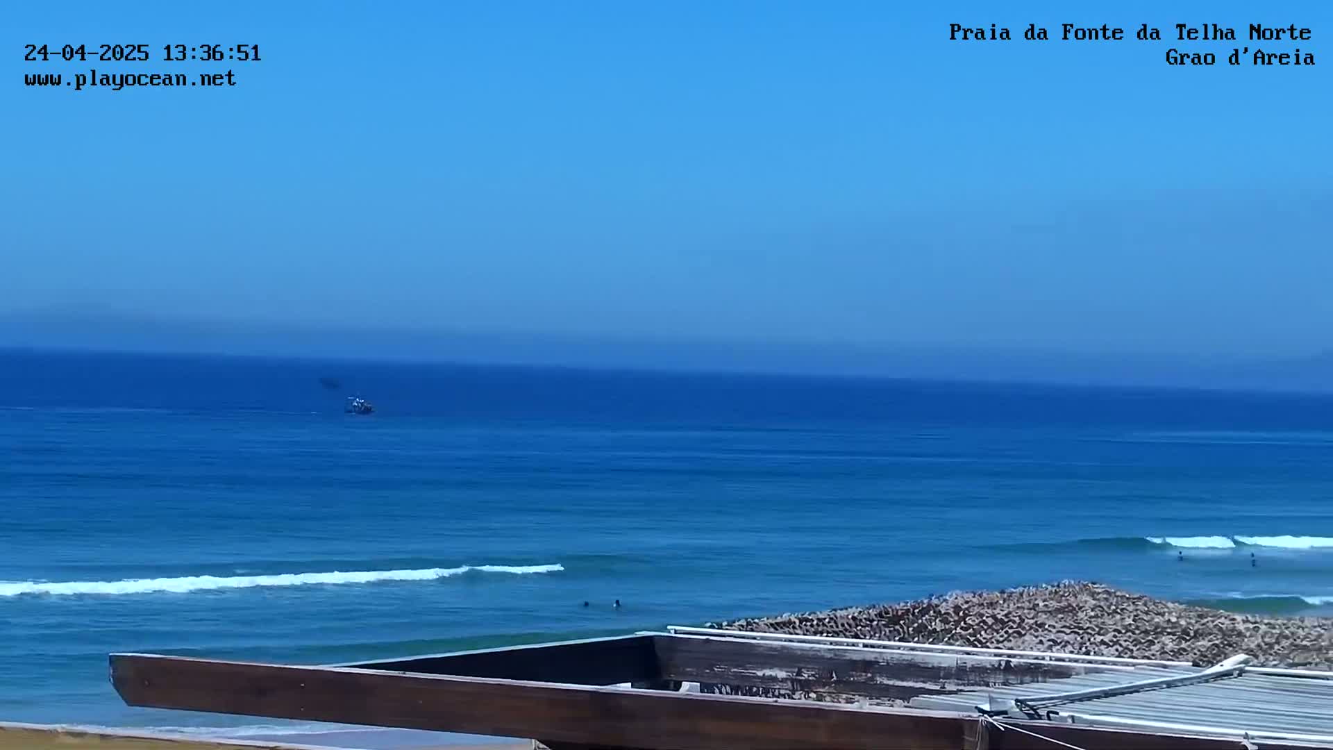 A mostly clear blue sky overlooks a tranquil ocean with small waves, a distant boat, and a few people in the water near a rocky shore and a wooden structure.