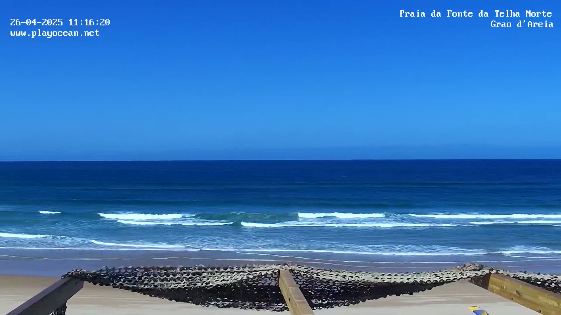 A sandy beach with a clear blue ocean and small waves under a bright blue sky is partially visible beyond a dark-colored protective barrier.