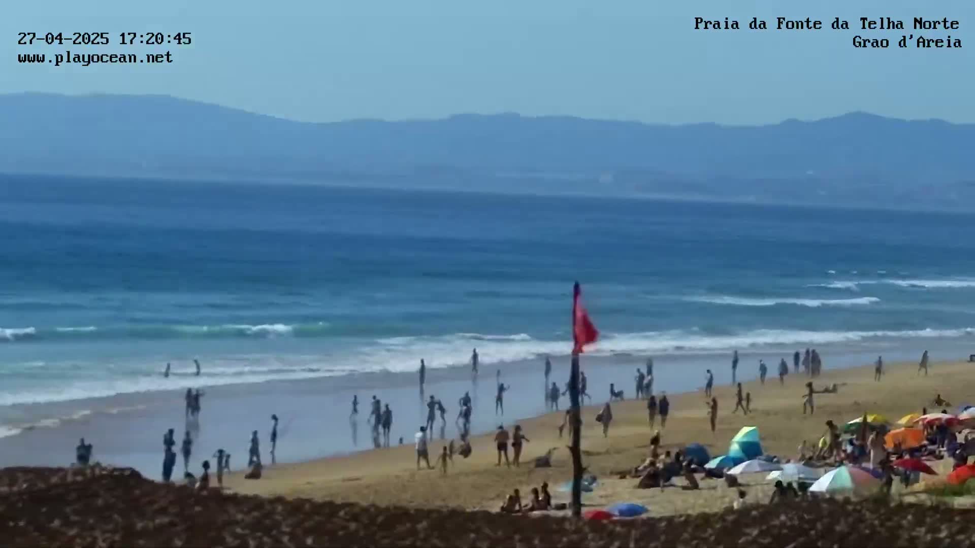A sandy beach with many people enjoying the sun and ocean under a clear sky, with gently rolling waves.