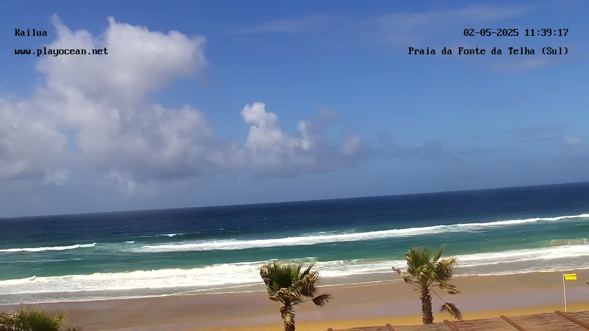 A partly cloudy day shows a wide sandy beach with gently rolling waves and a few palm trees.
