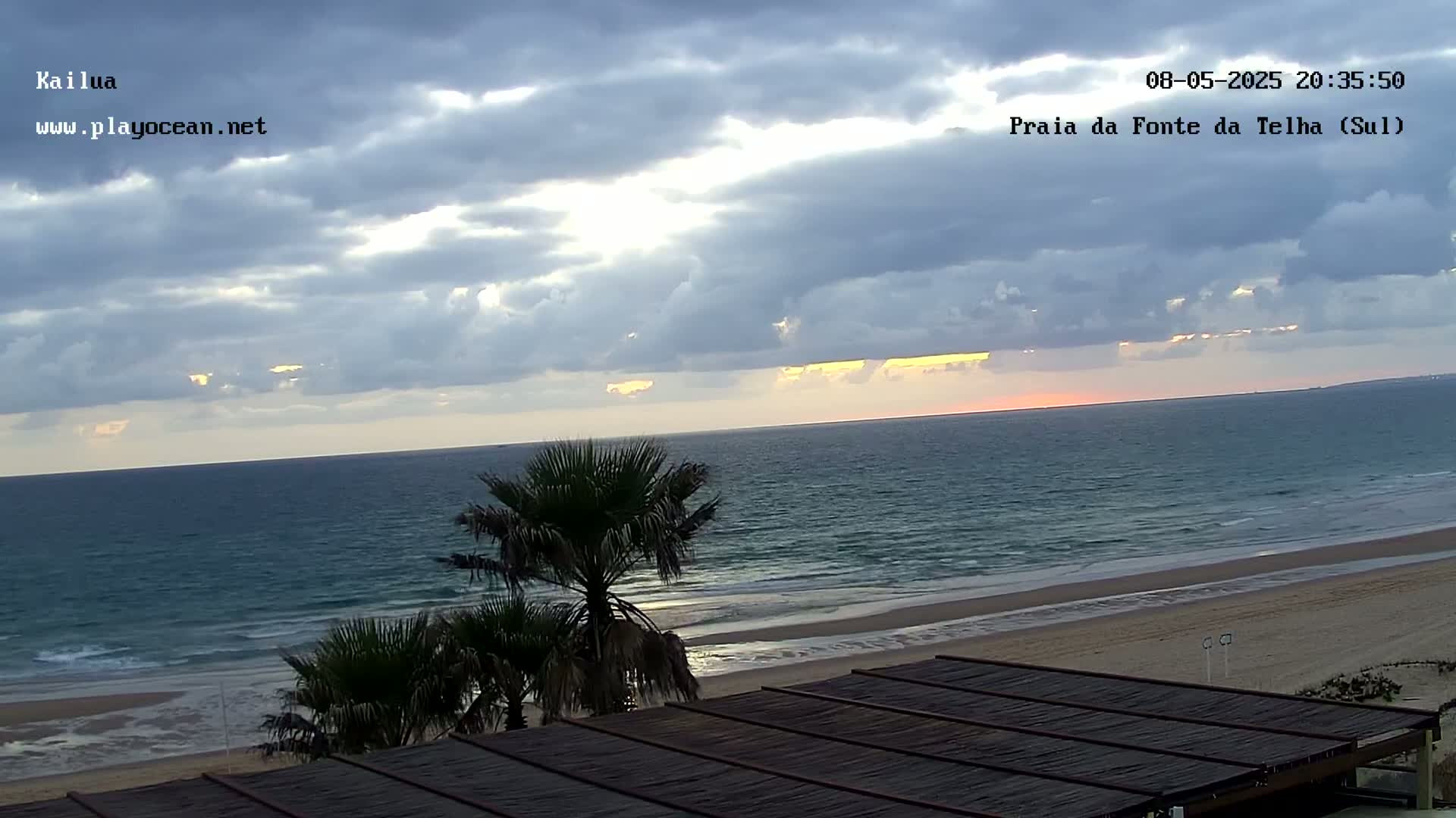 A partly cloudy sunset over a calm ocean and sandy beach, viewed from a slatted wooden structure with palm trees in the foreground.