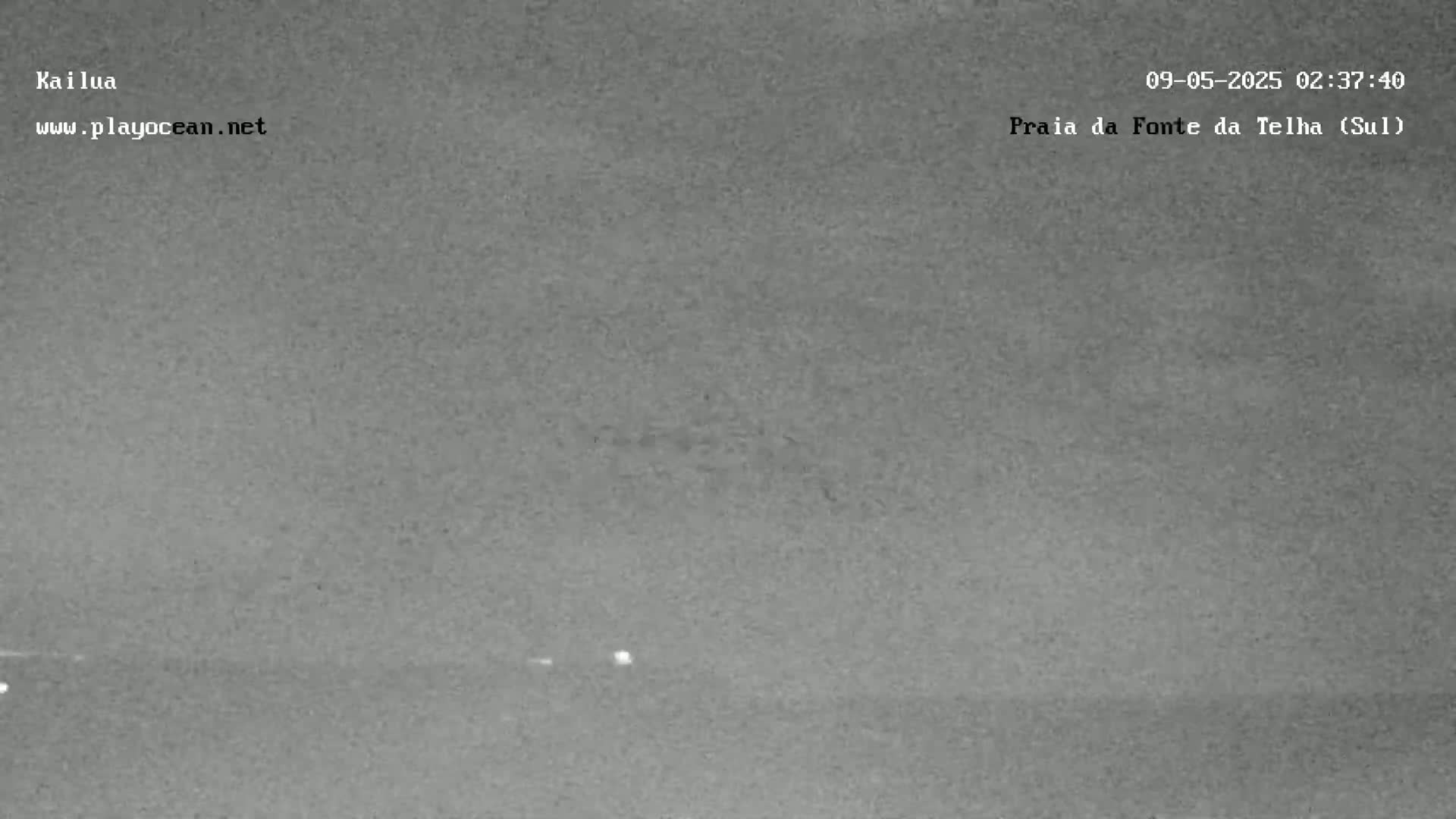 A nighttime, low-light image shows a dark, mostly featureless landscape with a few faint lights visible in the distance.