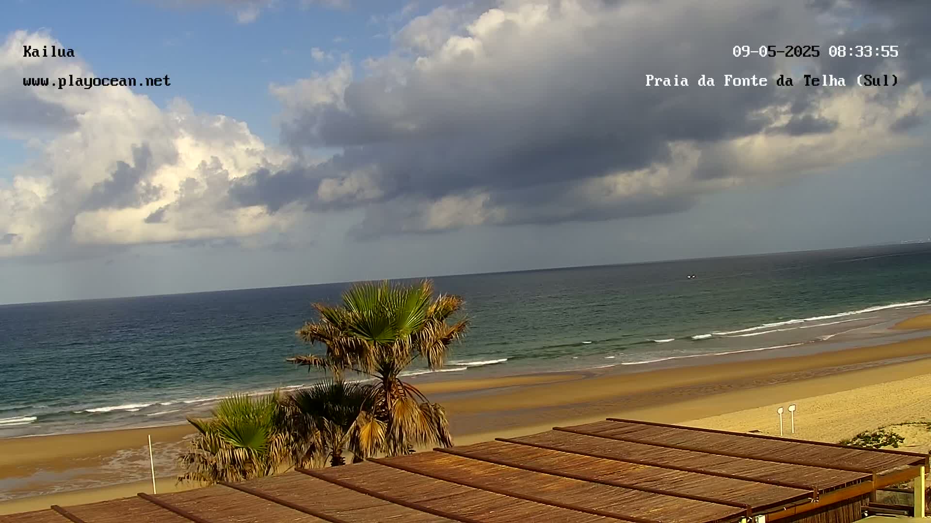 A partly cloudy sky overlooks a sandy beach and calm ocean, with palm trees in the foreground and a slatted wooden structure below.