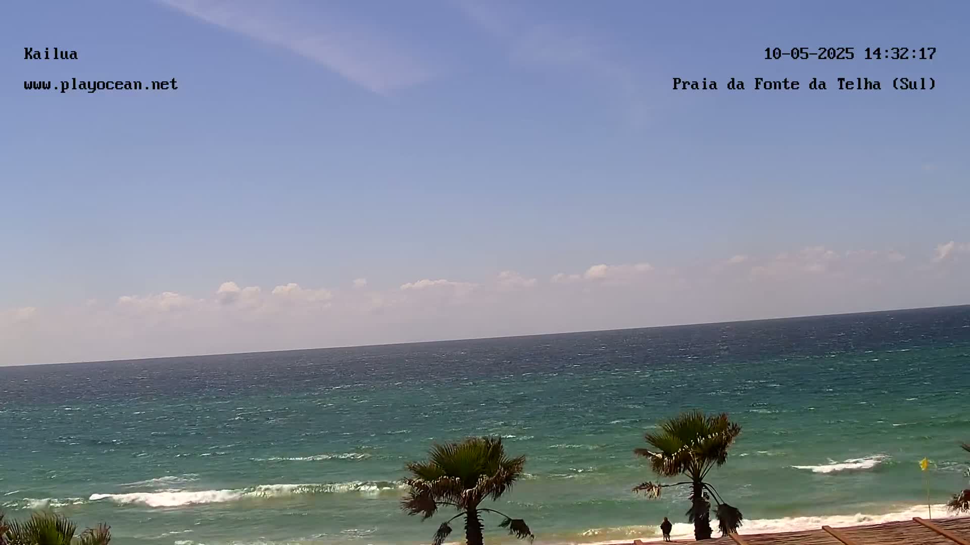 A partly cloudy day shows a wide expanse of teal ocean with small waves lapping a sandy shore, where palm trees stand near a wooden structure.