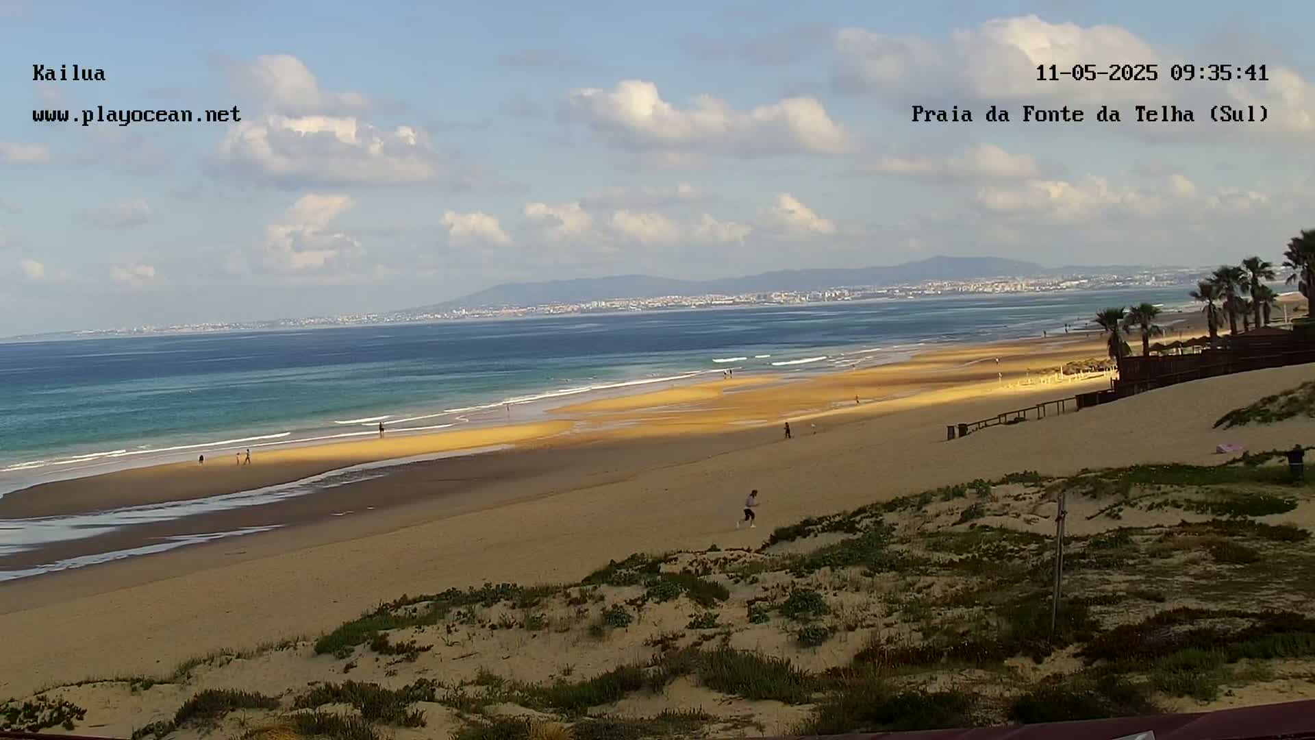 A long sandy beach stretches to a calm ocean under a partly cloudy sky, with a distant coastline visible.