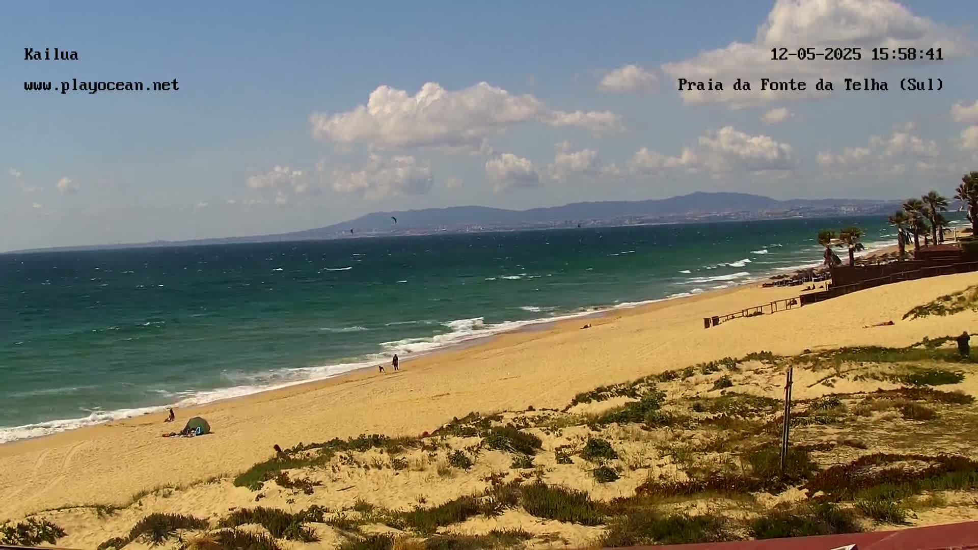 A partly cloudy day at a beach shows a person walking a dog on a wide sandy beach, with a few other people scattered about, and a somewhat choppy ocean with small waves.