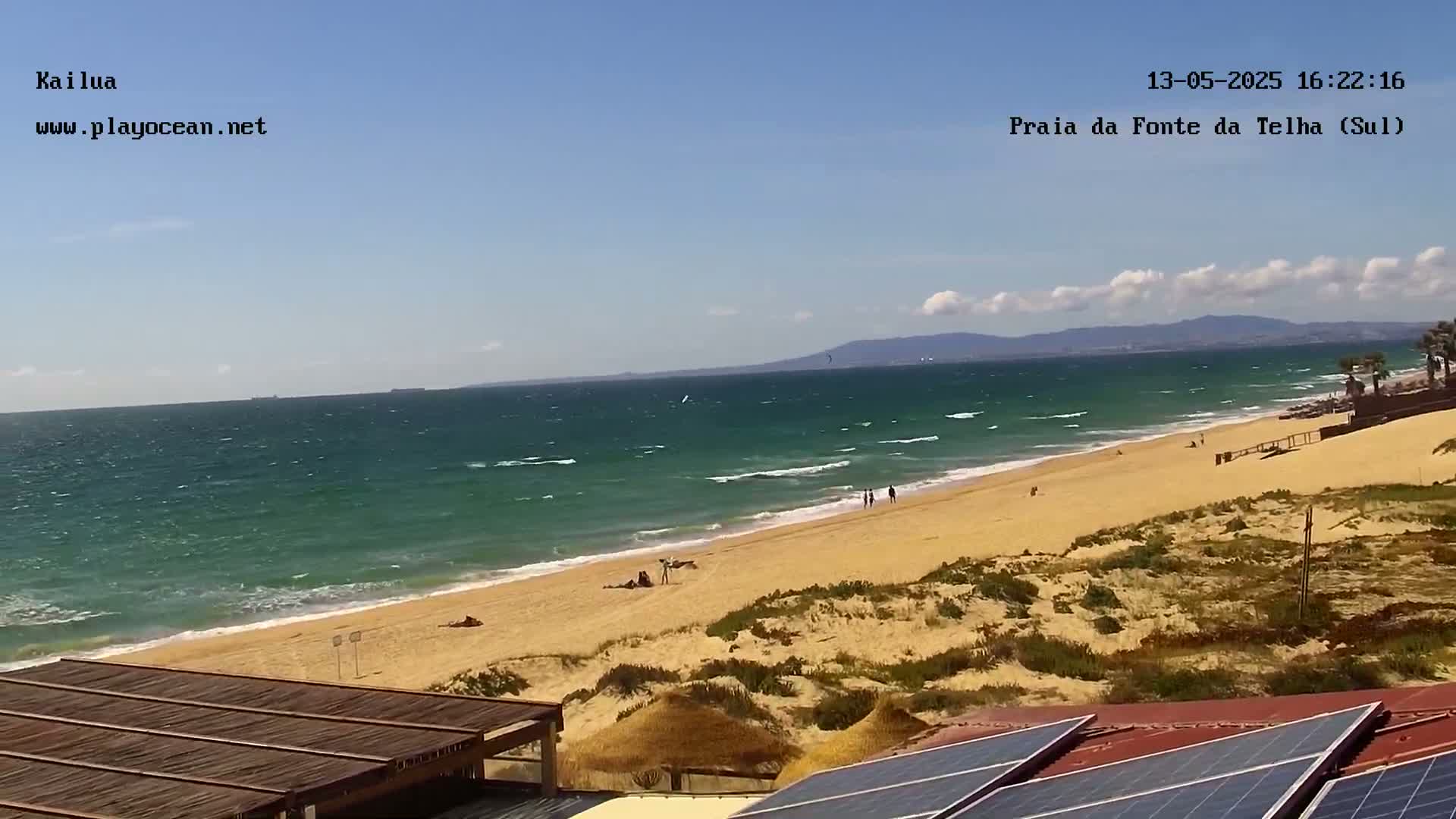 A sandy beach with a few people on it stretches along a teal-green ocean under a mostly sunny sky with some clouds.