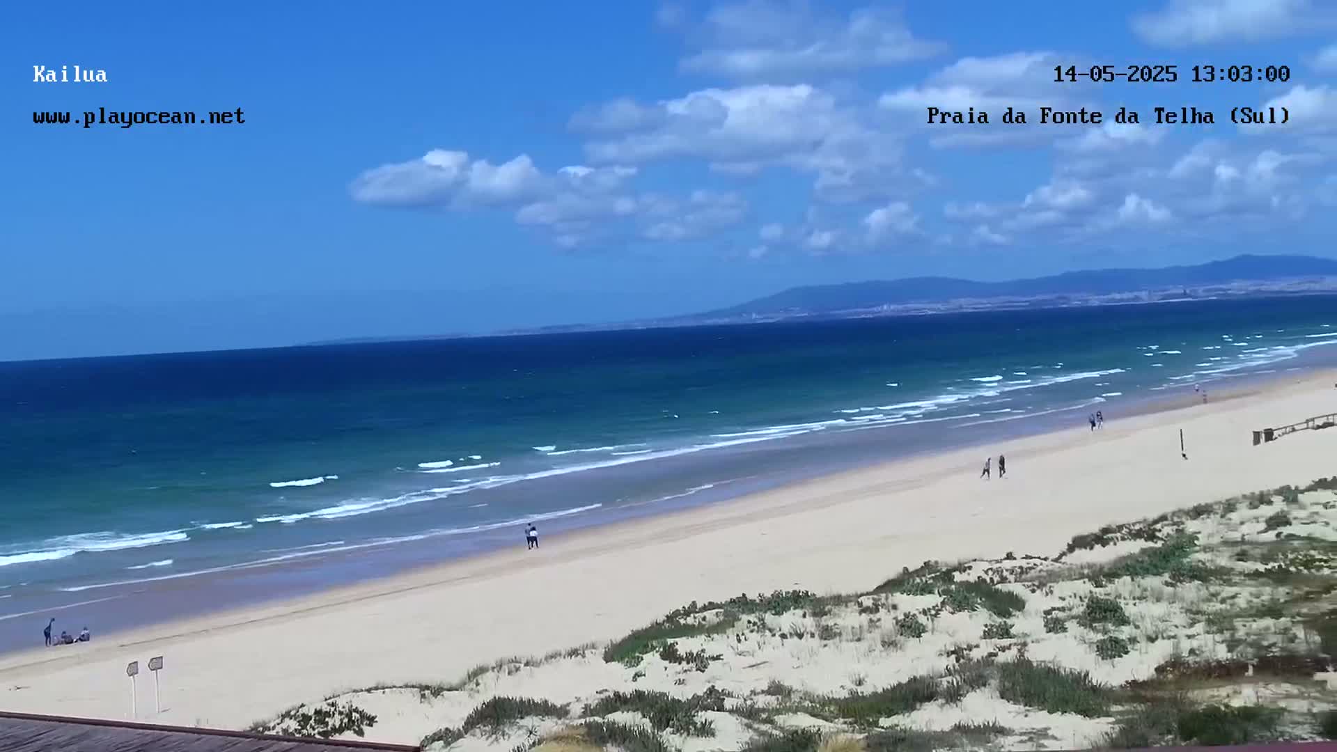 A sandy beach with a few people on it stretches out to a calm ocean under a mostly sunny sky.